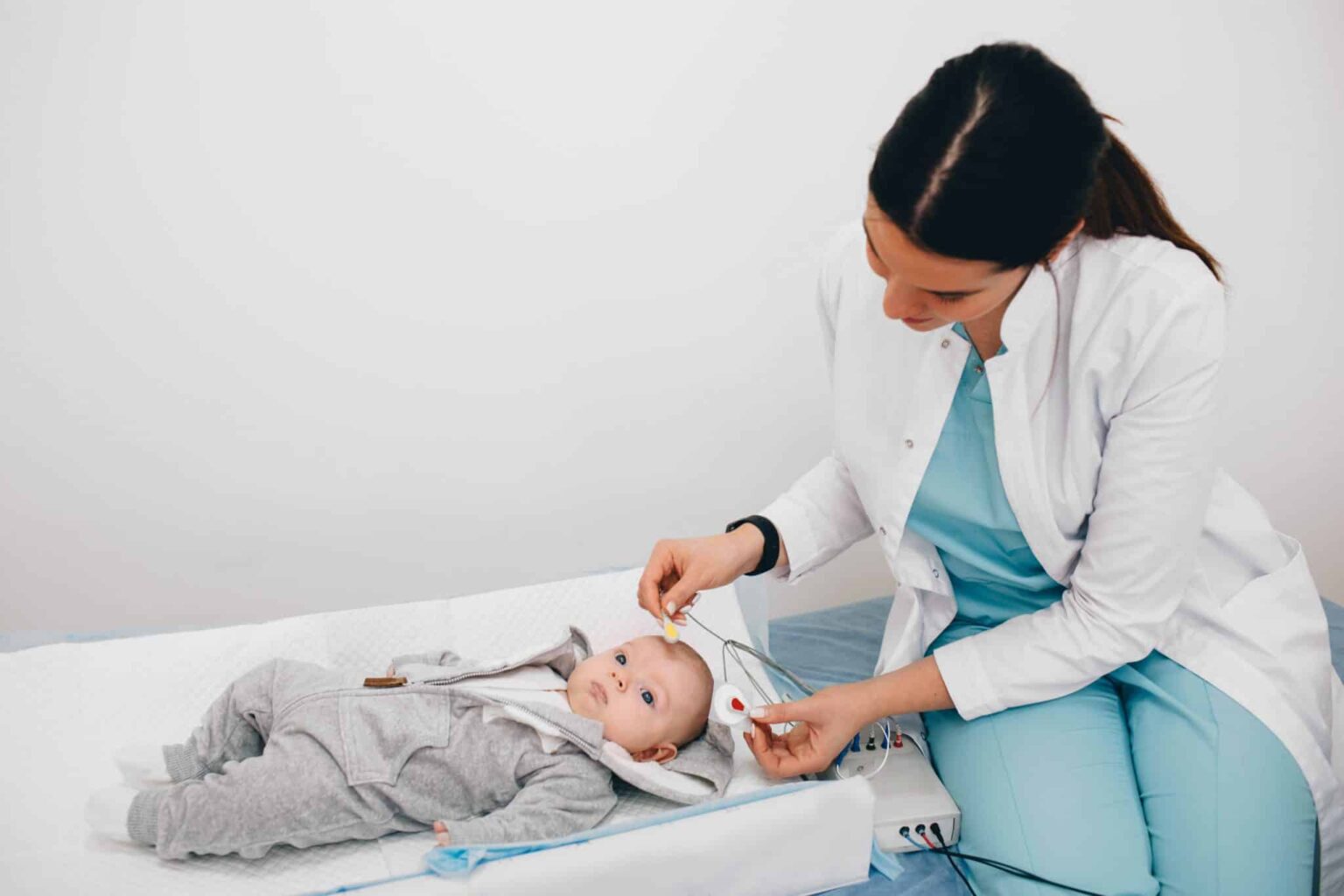 Baby in a hospital setting getting small electrodes placed on its head by a doctor who is leaning over it on a bench next to the small table the baby is lying on