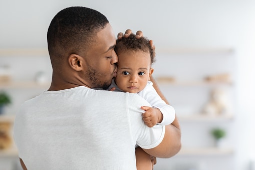 Man holding baby against his chest and kissing it on the cheek. 